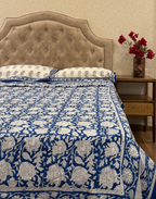 Bed with blue and white floral patterned bedding, brown upholstered headboard, and nightstand with red flowers.